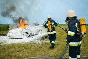 Firefighters tackle a blazing car using foam to extinguish the flames in an outdoor setting.