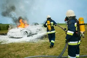 Firefighters tackle a blazing car using foam to extinguish the flames in an outdoor setting.