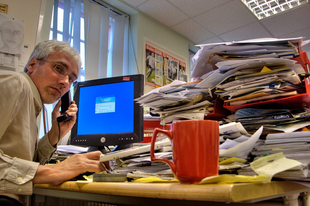A real estate agent working late at a desk buried in paperwork and a laptop